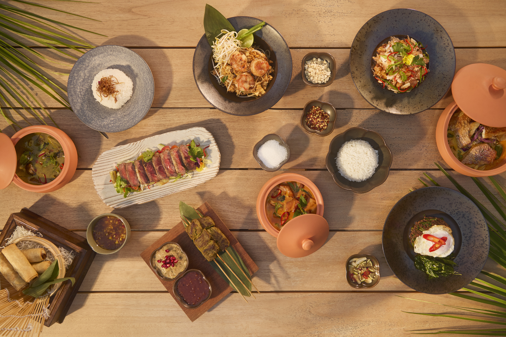 Top-down view of a wooden table set with a variety of vibrant Thai dishes, including jasmine rice, grilled skewers, spring rolls, sliced seared meat, shrimp pad Thai, red and green curries in clay pots, garnishes like peanuts and chili flakes, and dipping sauces, all surrounded by tropical leaves.